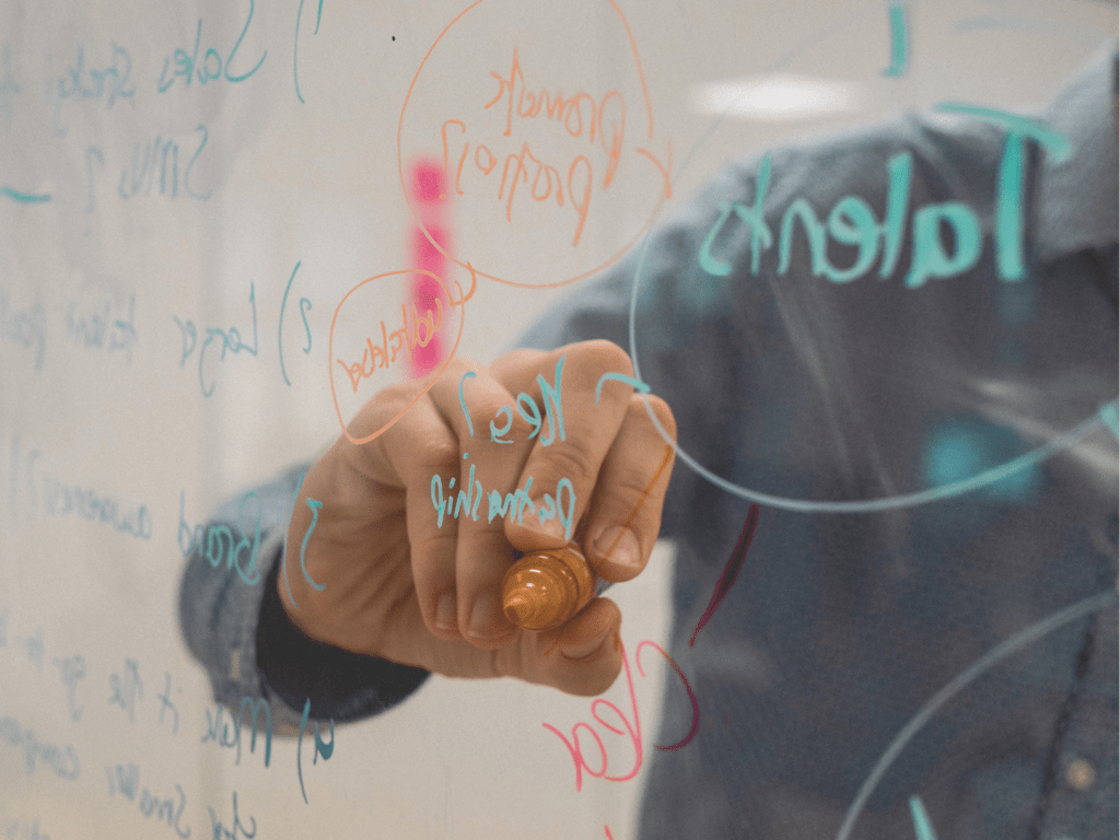 eine Person zeichnet Formeln mit einem bunten Stift an eine durchsichtige Tafel