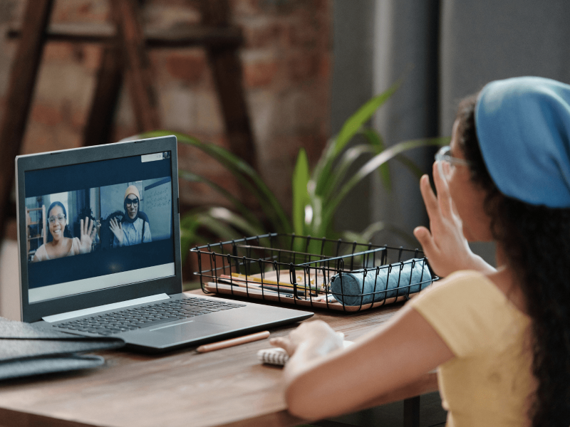eine junge Frau winkt mit der Hand vor ihrem Laptop den Menschen im Meeting zu