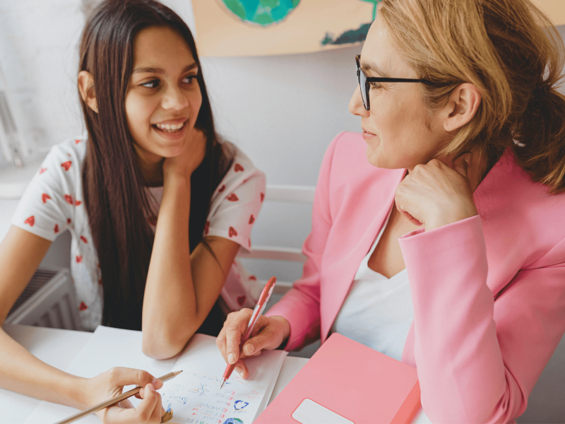 eine Schülerin und eine Lehrkraft sitzen gemeinsam am Schreibtisch und lächeln sich an