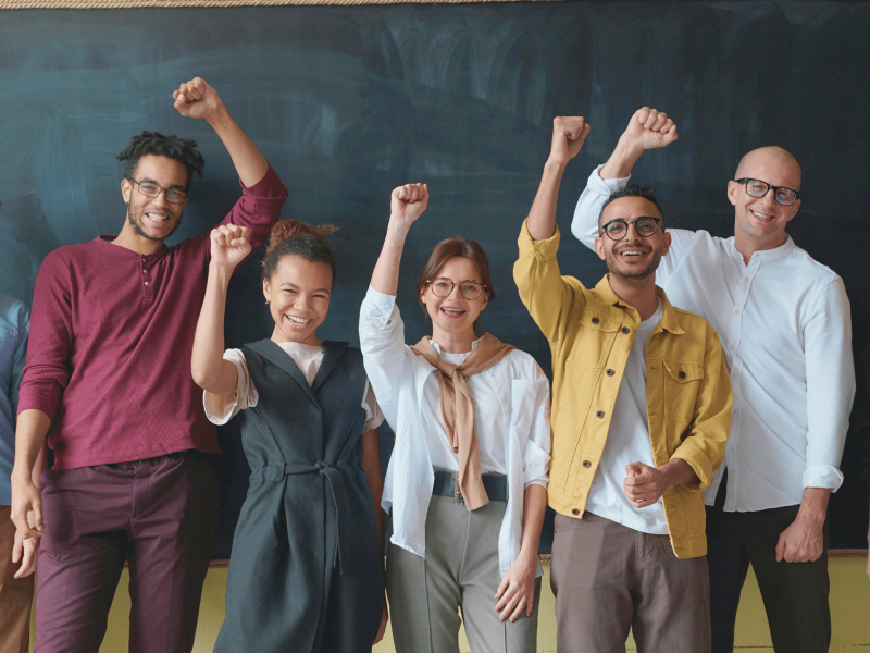 Diverse Lehrkräfte stehen vor einer Tafel und heben lächelnd und selbstbewusst die Faust